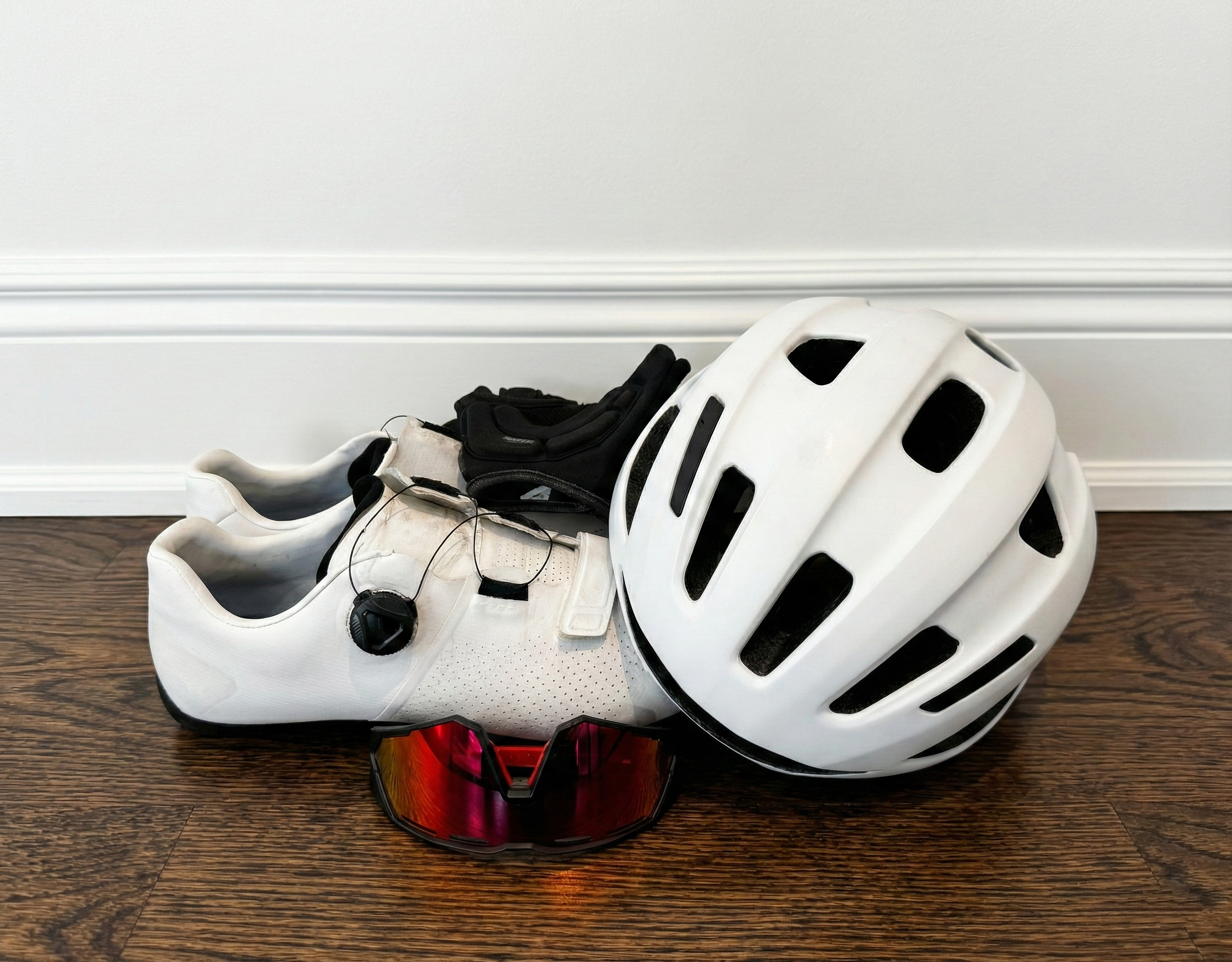 a pile of cycling accessories sits on a hardwood floor. There is a white helmet, black gloves, white cycling shoes, and red sunglasses.